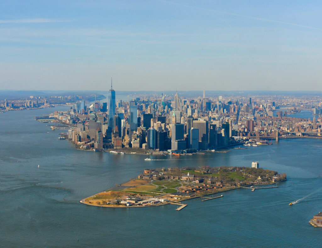 Aerial view of Governors Island with its lawns, paths and historic buildings, and the Lower Manhattan skyline across the water