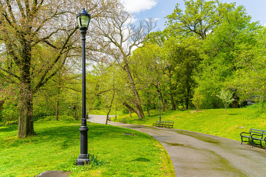 An empty curving path through Prospect Park in spring, with an old-style lamp post, benches and fresh green trees