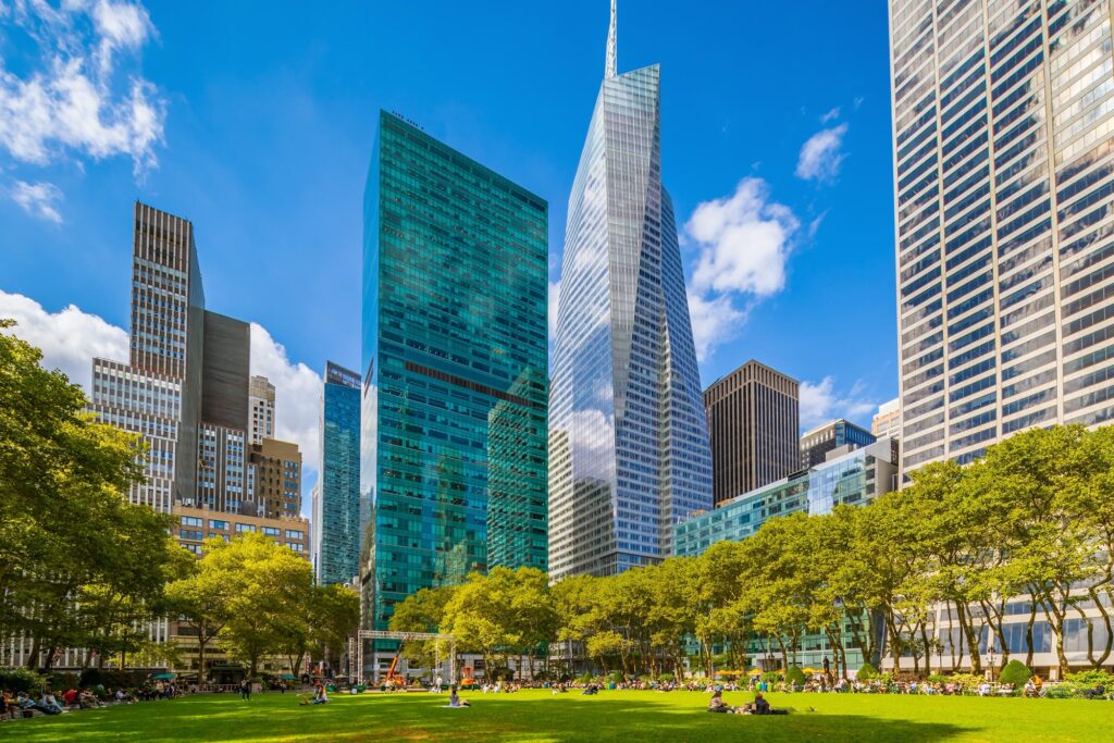 People sitting on the lawn of Bryant Park on a sunny day, ringed by plane trees and Midtown skyscrapers