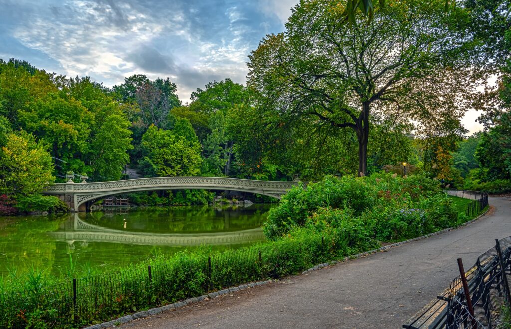 Bow Bridge in Central Park reflected in the Lake on a late-summer morning, with a curving path and benches