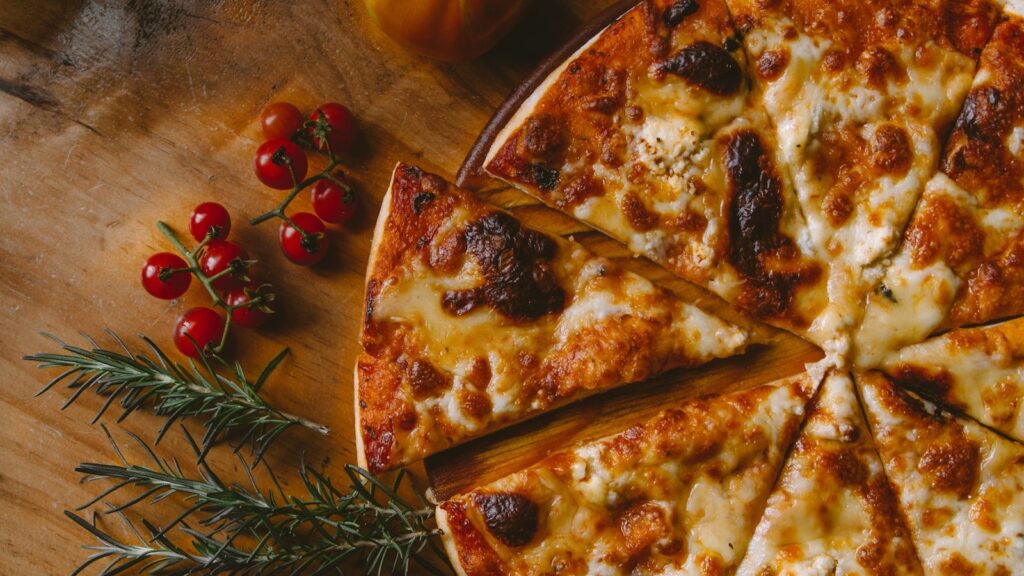 A freshly sliced New York-style cheese pizza on a wooden board surrounded by cherry tomatoes and rosemary