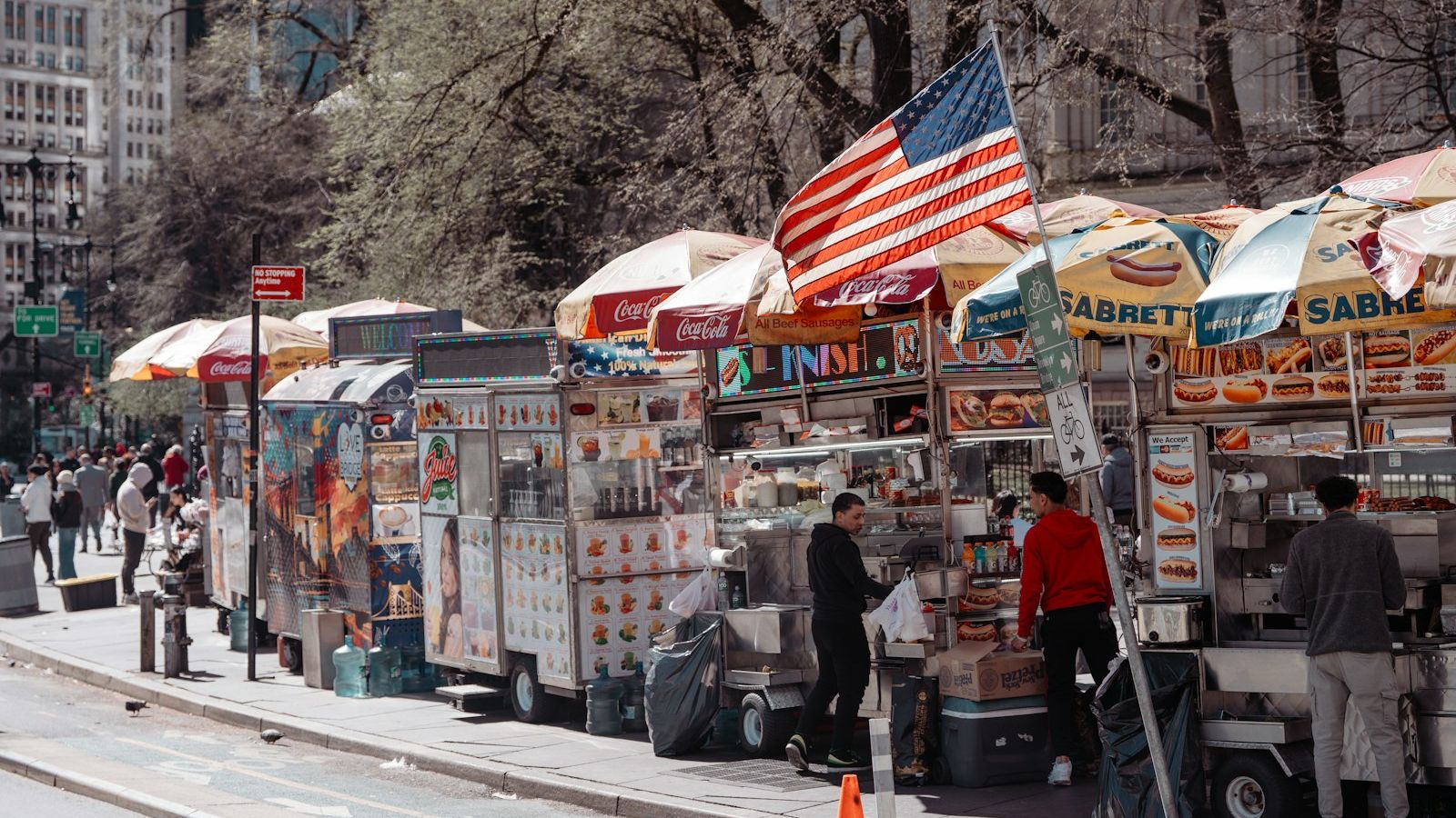 Food vendor carts lined up on a Manhattan sidewalk with an American flag flying above them on a spring day in New York City