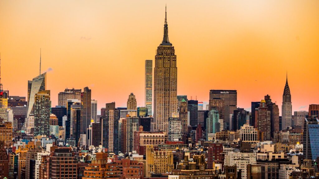 The Manhattan skyline at golden hour, with the Empire State Building and Chrysler Building rising above Midtown — the iconic view from New York Harbor and Governors Island