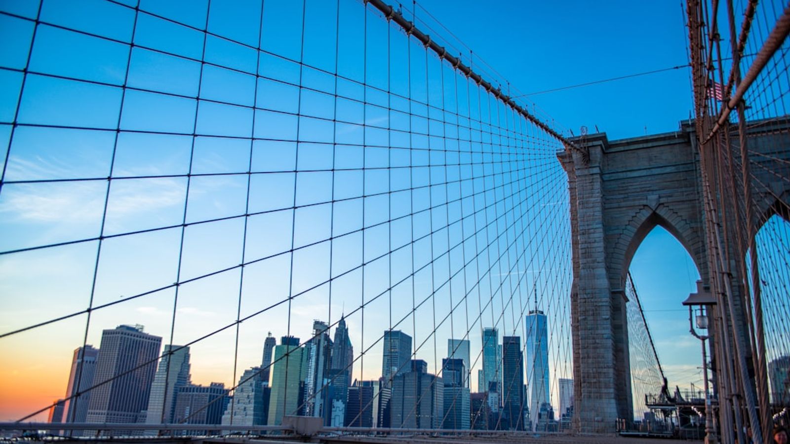 View through Brooklyn Bridge cables toward the Gothic stone tower and Manhattan skyline at sunset
