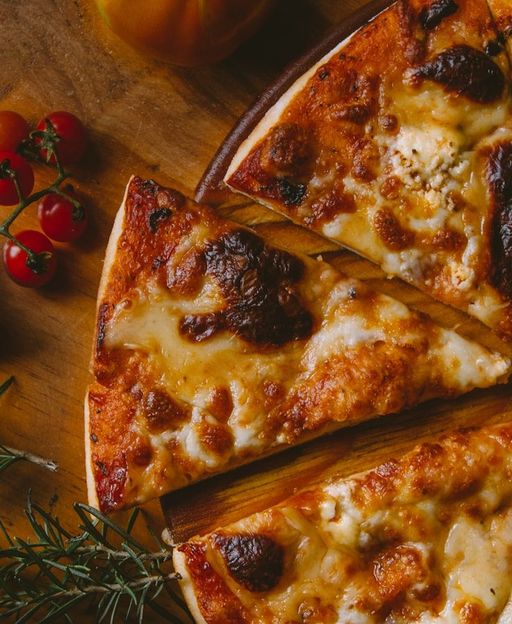 A freshly sliced New York-style cheese pizza on a wooden board surrounded by cherry tomatoes and rosemary