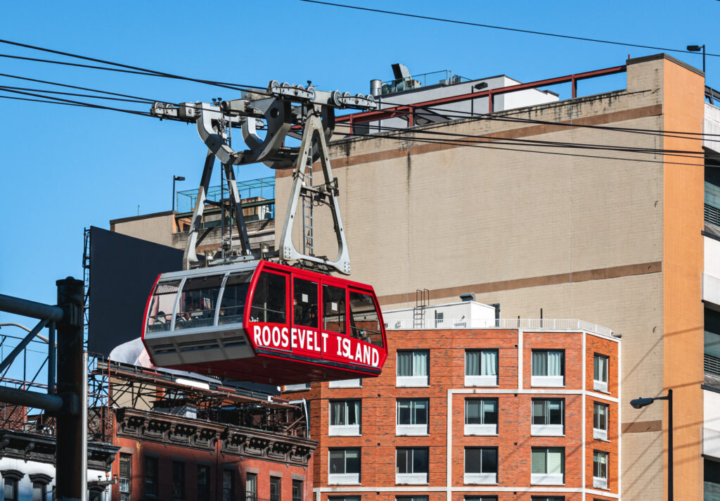A red Roosevelt Island tram cabin gliding on its cables past brick and beige buildings in Midtown East under a blue sky