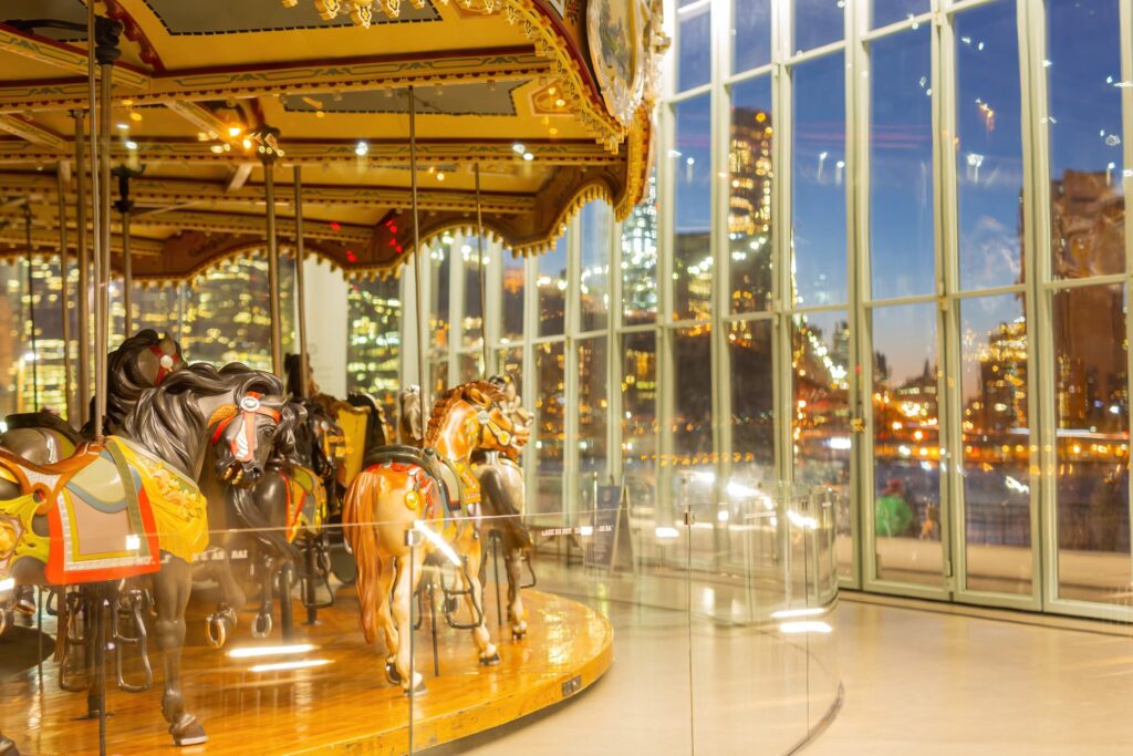 Painted horses on Jane’s Carousel inside its glass pavilion at dusk, with the lit Manhattan skyline beyond the windows