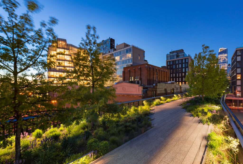 The High Line walkway at dusk, lit planting beds and young trees beside the path, with Chelsea buildings glowing behind