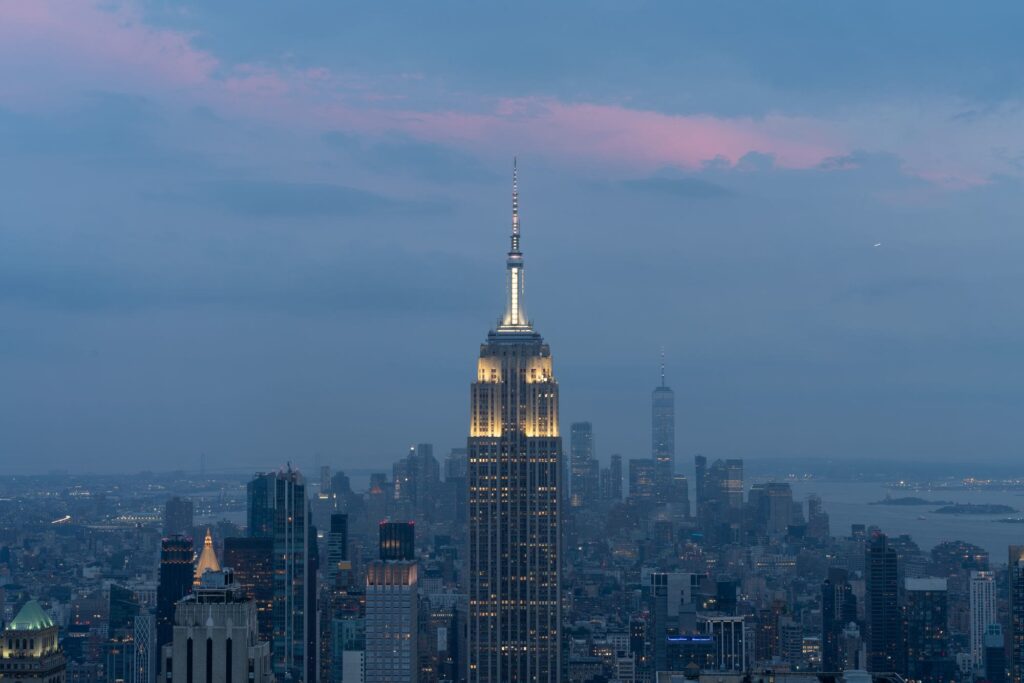 The Empire State Building lit up at dusk above Midtown, with One World Trade Center and the harbor in the distance under pink-tinged clouds