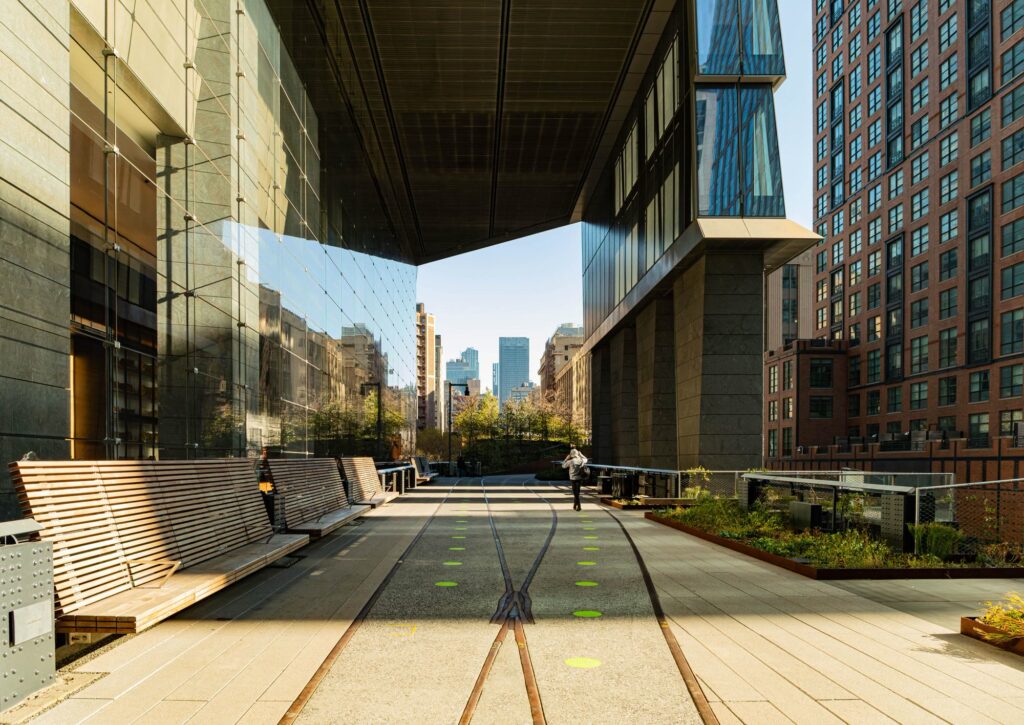 Old rails set into the paving of the High Line beneath an overhanging glass tower at Hudson Yards, with long wooden benches and a planting bed