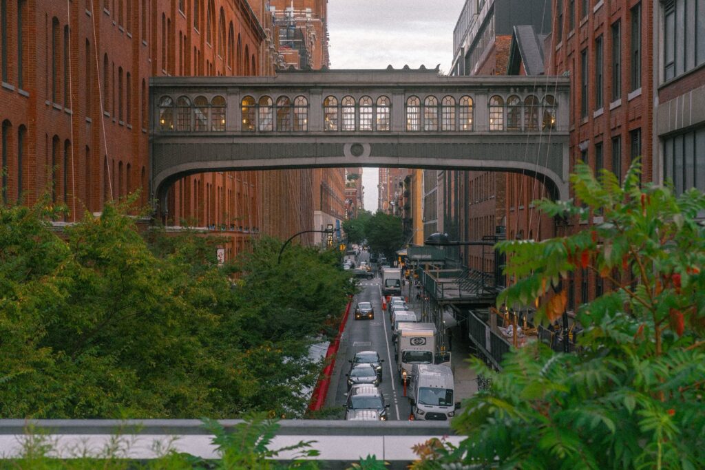 Red-brick former factory buildings joined by an arched stone skybridge, seen from the planted edge of the High Line above a Chelsea street