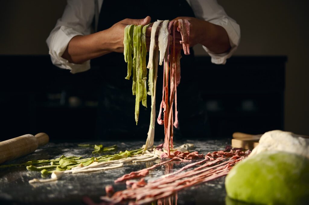A chef lifting strips of green, white and red handmade pasta over a floured counter