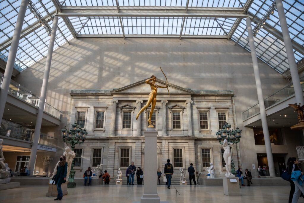 A gilded statue of Diana with her bow in the glass-roofed American Wing courtyard at the Met, with marble statues and visitors around it