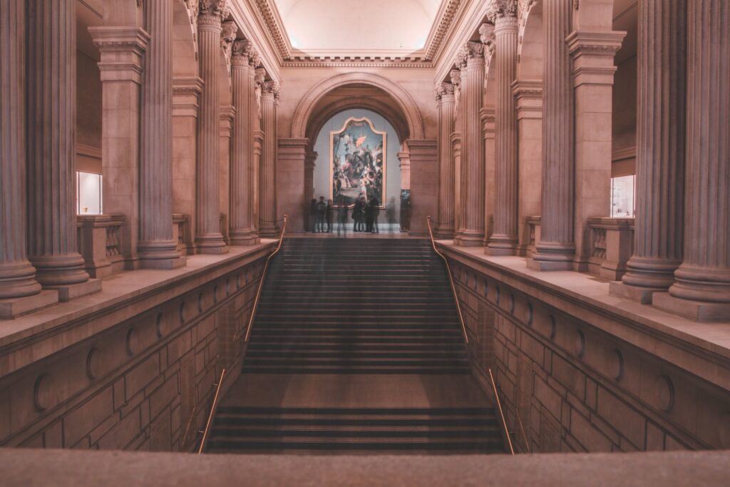The grand staircase rising from the Great Hall of the Metropolitan Museum of Art, lined with stone columns, with a large painting at the top