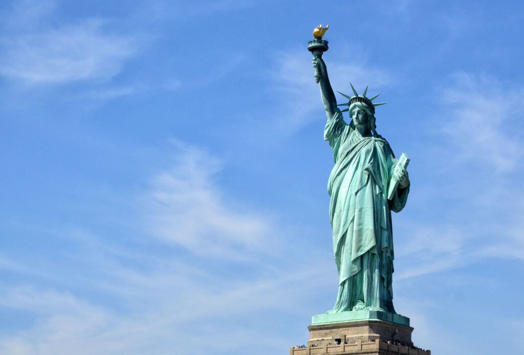 The Statue of Liberty on its pedestal on Liberty Island under a blue sky, with visitors on the pedestal balcony