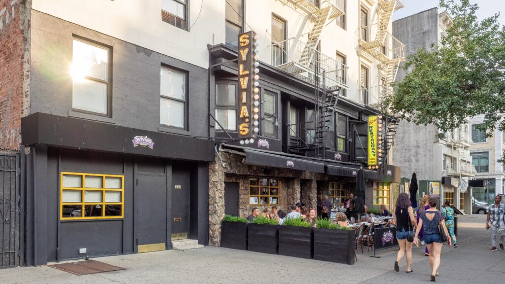 Sylvia's Restaurant on Lenox Avenue in Harlem, with its lit vertical sign and diners at sidewalk tables