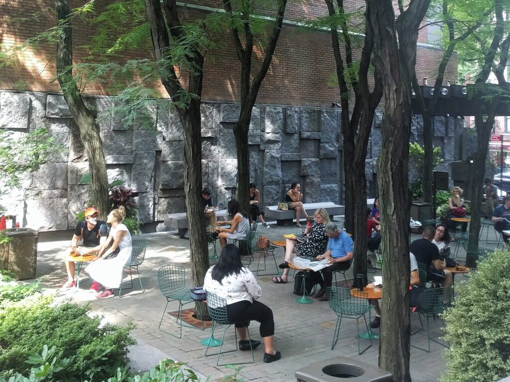 People reading and eating lunch at small tables under the trees in Greenacre Park, seen from the raised platform, with the rough granite wall behind