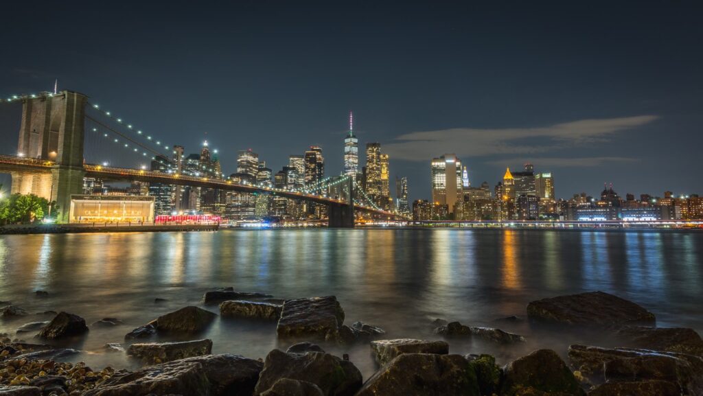 The Brooklyn Bridge and the lit Lower Manhattan skyline at night, seen across the East River from the rocks in Brooklyn Bridge Park, with Jane's Carousel glowing by the water