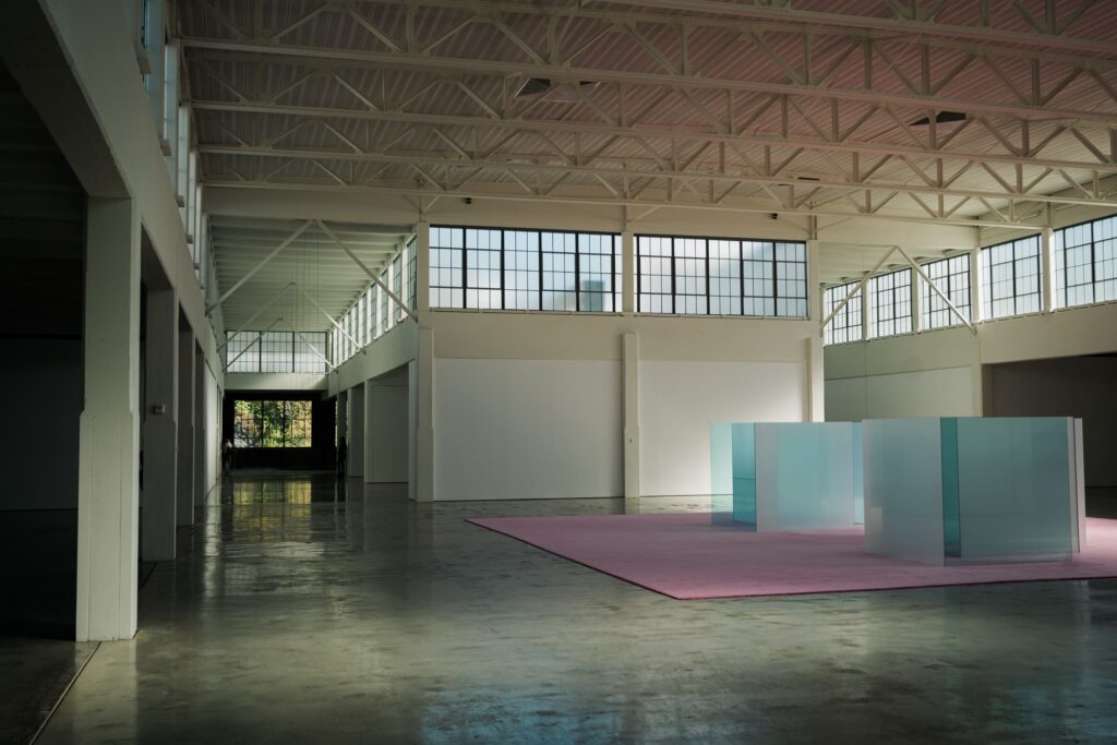 Sunlit gallery inside Dia Beacon, with a glass-panel sculpture on a pink carpet under factory skylights