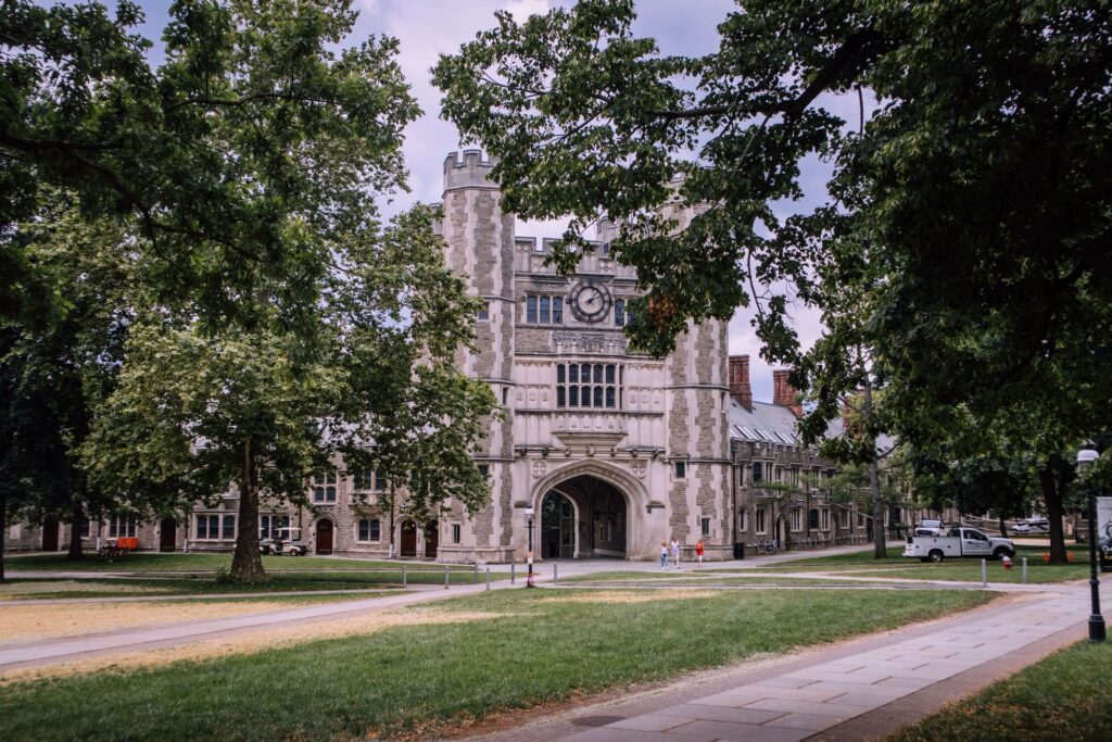 Gothic stone gatehouse with a clock tower and arched passage on the Princeton University campus, framed by trees