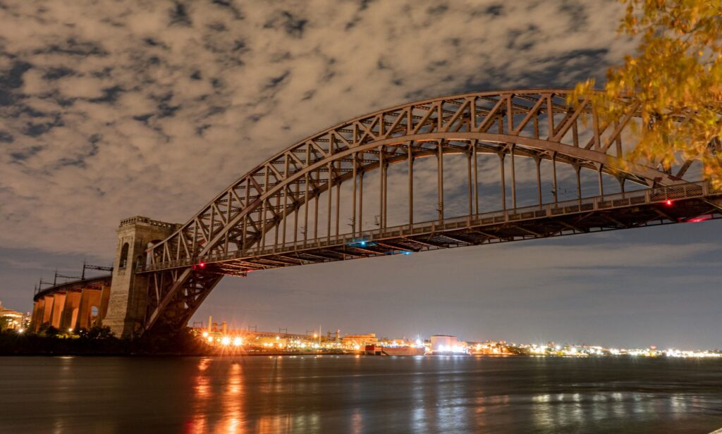 Hell Gate Bridge lit at night over the East River beside Astoria, Queens