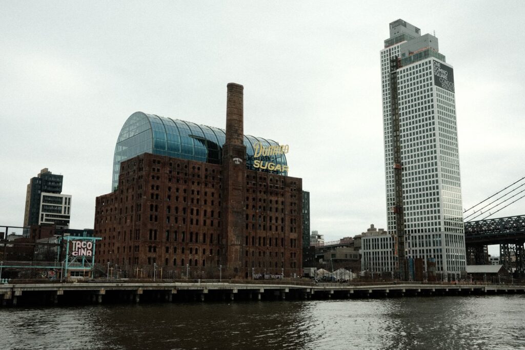 The old Domino Sugar refinery and the Domino Park waterfront in Williamsburg seen from the East River on a gray day, with the Williamsburg Bridge at the right edge