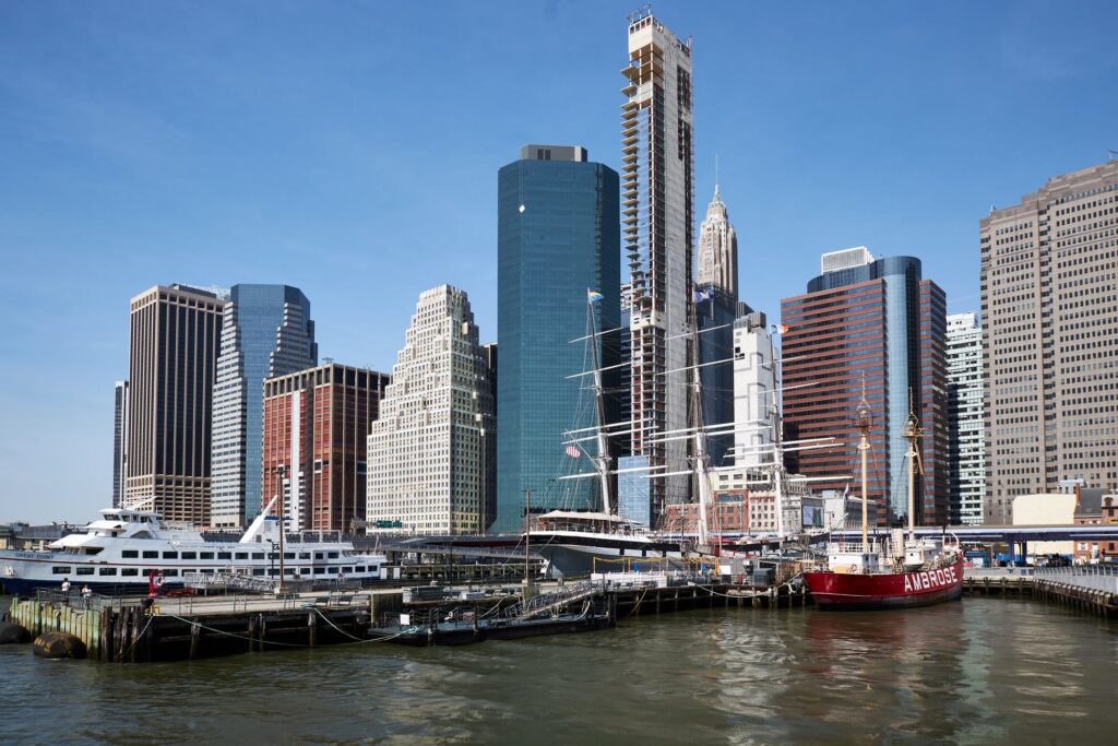 The South Street Seaport piers by day, with the tall ship Wavertree and the red Ambrose lightship moored in front of Lower Manhattan towers