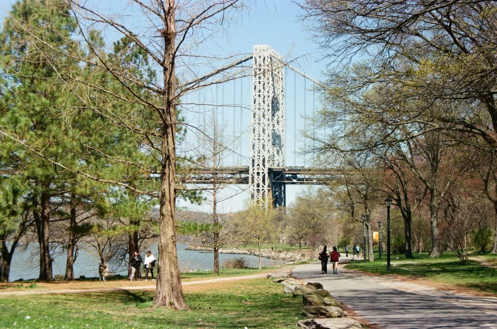Walkers on a tree-lined riverside path beside the Hudson on a spring day, with a tower of the George Washington Bridge rising ahead