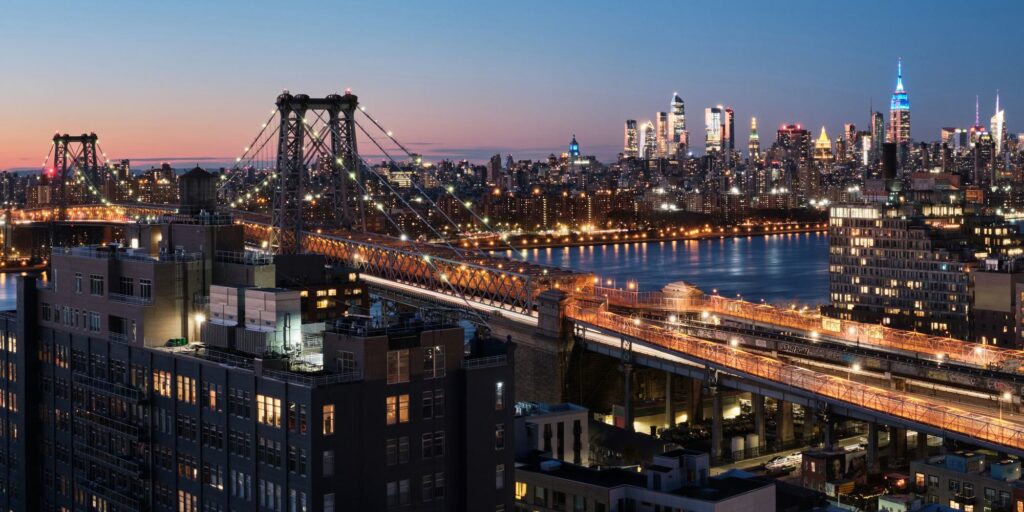 The Williamsburg Bridge at dusk from above Brooklyn, lights along its cables and its orange-lit truss and walkway crossing the East River toward the Midtown Manhattan skyline