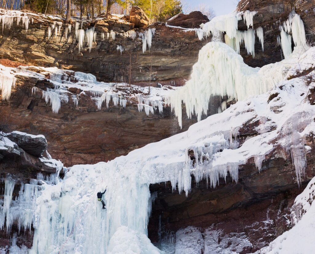 Kaaterskill Falls frozen into curtains of ice over red rock ledges in winter, with an ice climber on the lower tier