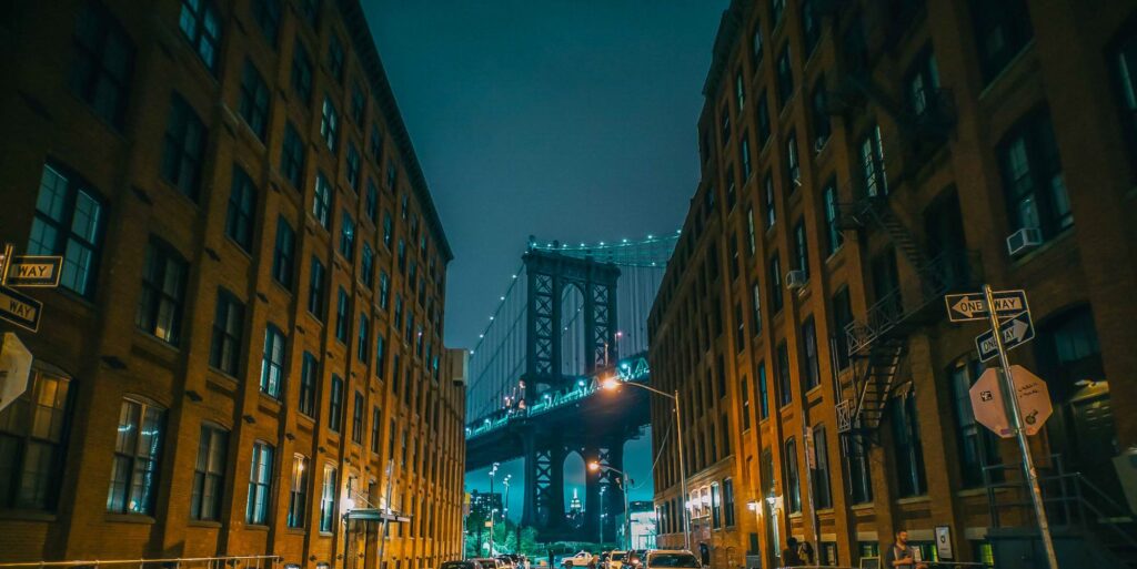 Washington Street in DUMBO at night, brick warehouses on both sides framing the lit Manhattan Bridge tower, with the Empire State Building visible through its base