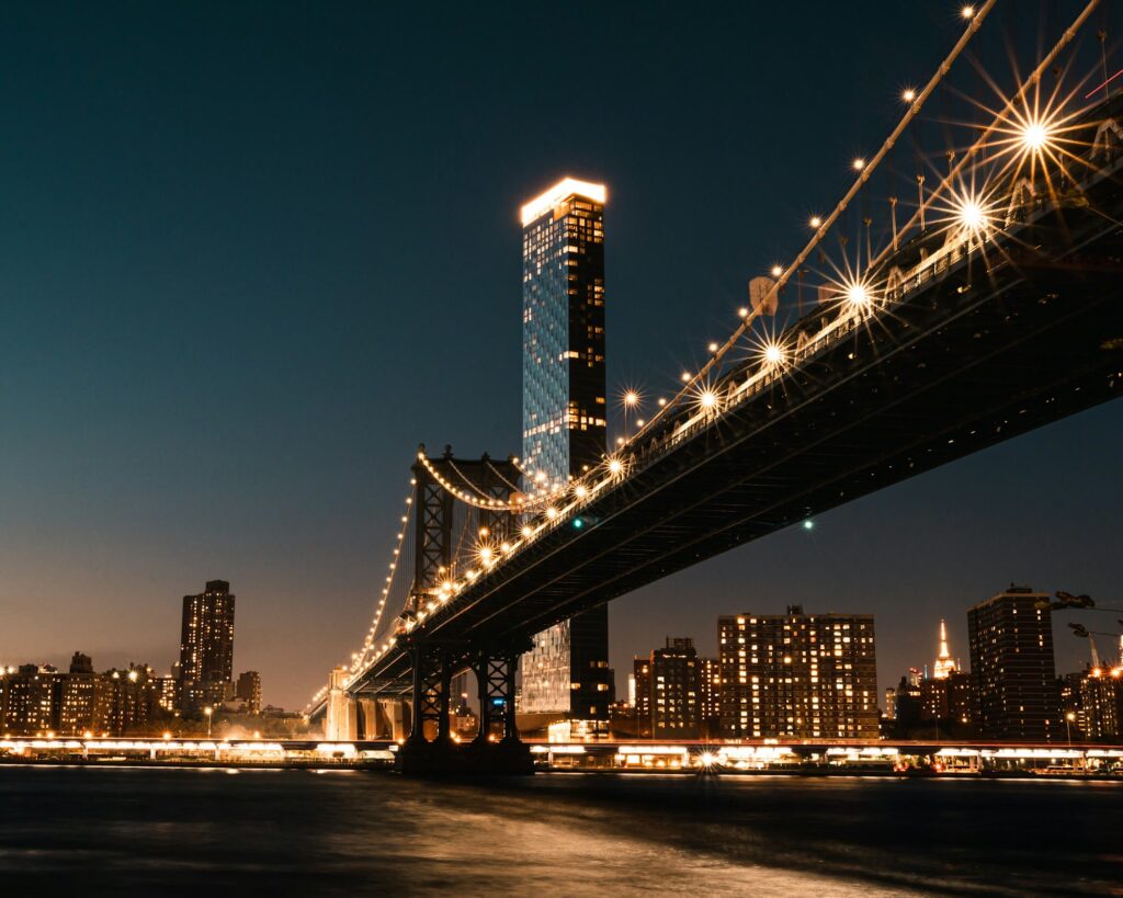 The Manhattan Bridge at night seen from the Brooklyn shore below it, lights strung along its cables and deck, with Manhattan apartment towers lit up beyond