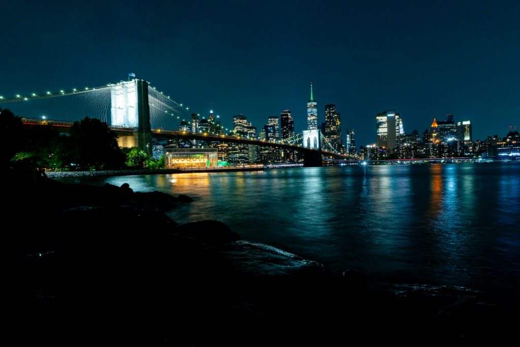 Brooklyn Bridge Park at night from the rocky shore, with Jane's Carousel lit up under the Brooklyn Bridge and the Lower Manhattan skyline across the river