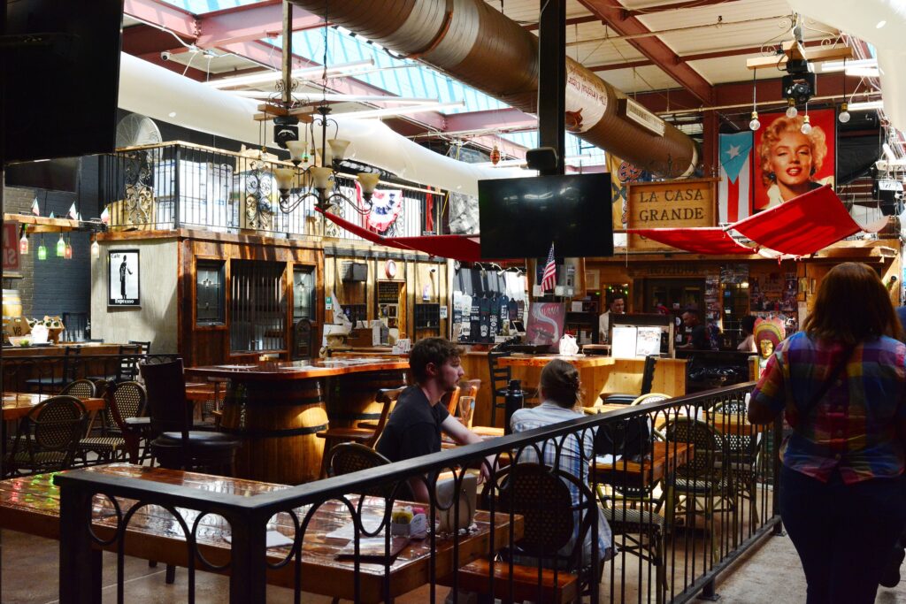 Diners at wooden tables inside the Arthur Avenue Retail Market, with the La Casa Grande cigar stand behind them