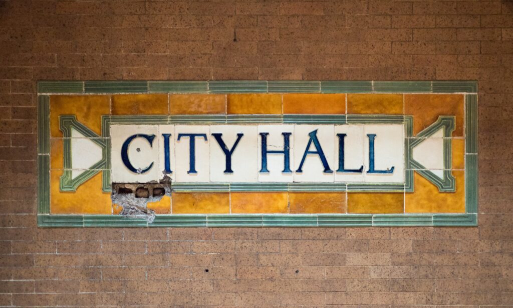The original glazed-tile CITY HALL name panel in green, gold and cream on the brick wall of the closed City Hall subway station