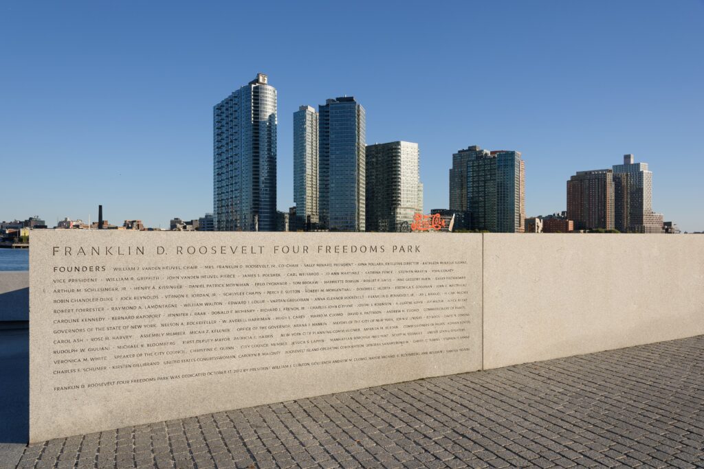 An engraved granite wall reading Franklin D. Roosevelt Four Freedoms Park, with the East River and Long Island City towers behind