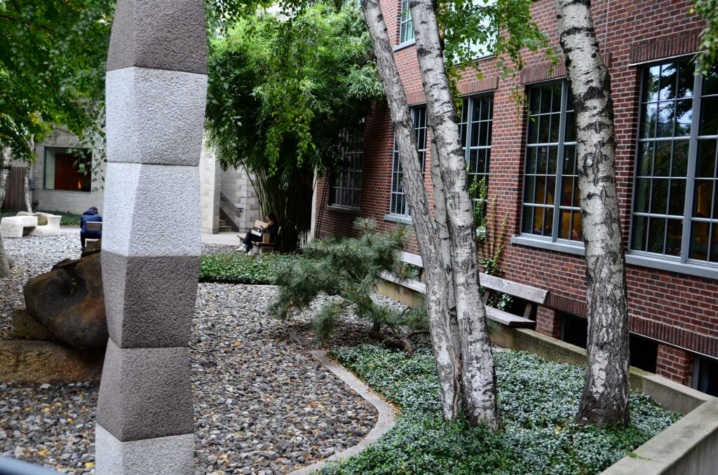 A stacked stone column, birch trees and gravel beds in the sculpture garden of the Noguchi Museum, beside the red-brick building