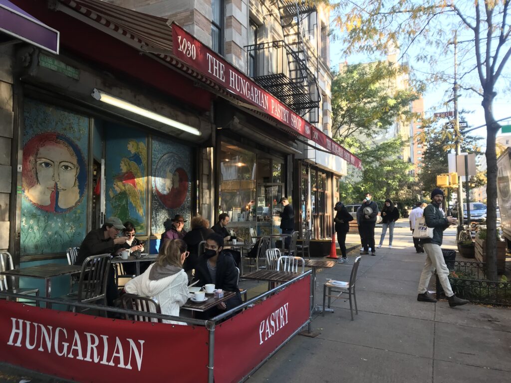 People drinking coffee at sidewalk tables under the red awning of The Hungarian Pastry Shop at 1030 Amsterdam Avenue
