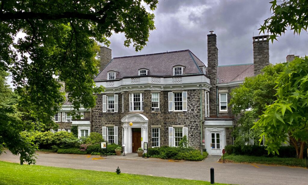 The stone front of Wave Hill House with white shutters and a white columned doorway, framed by green trees