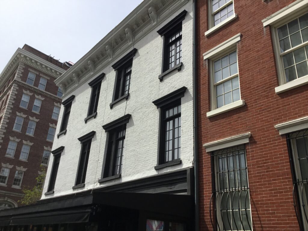 A white-painted brick townhouse with black window frames beside a red-brick row house on a West Village street in spring
