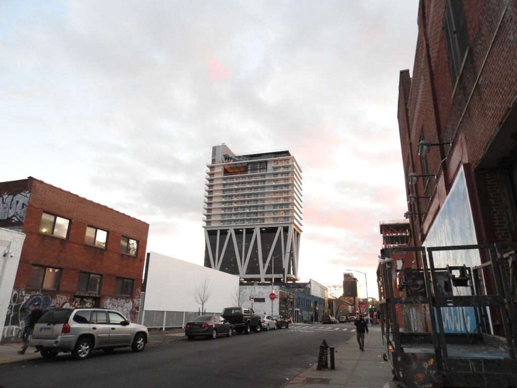 The William Vale hotel tower on its angled white stilts, rising above low brick buildings on a Williamsburg street at dusk