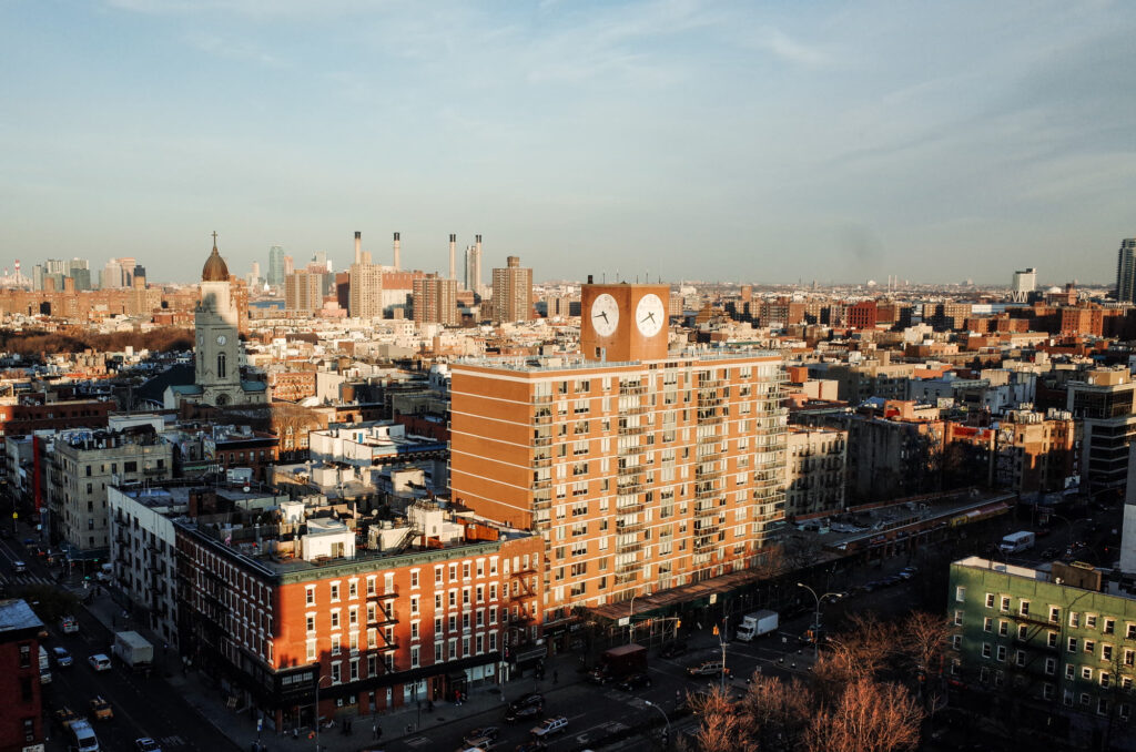 Elevated view over Lower East Side rooftops, a clock-topped apartment tower and a church dome in late-afternoon light