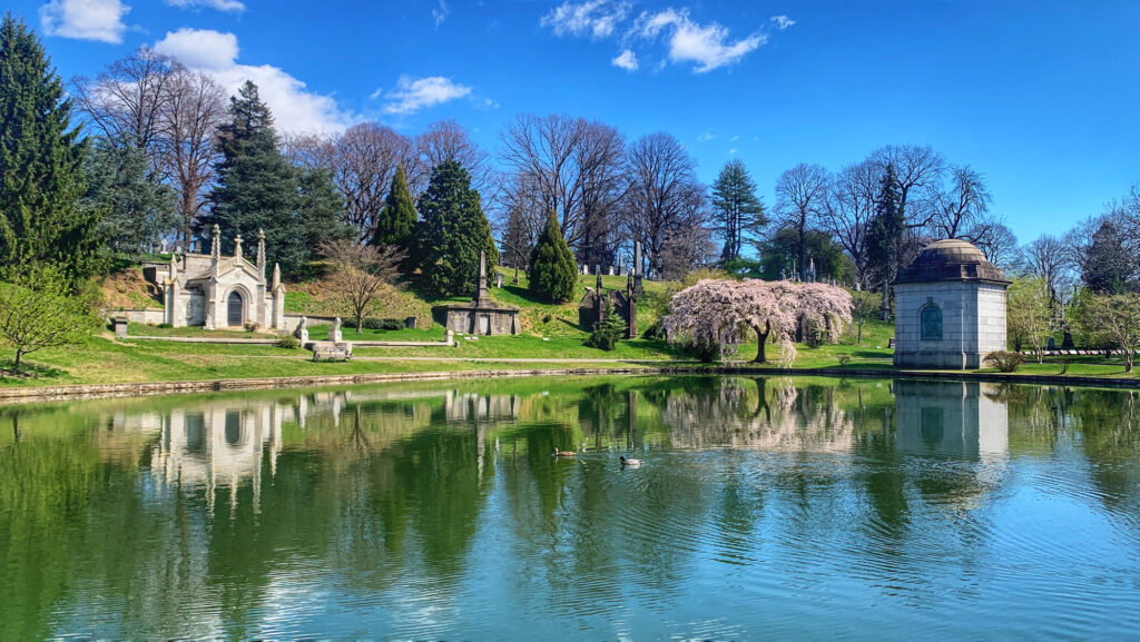 A still pond in Green-Wood Cemetery reflecting a Gothic mausoleum, a domed tomb and a pink weeping cherry tree in spring
