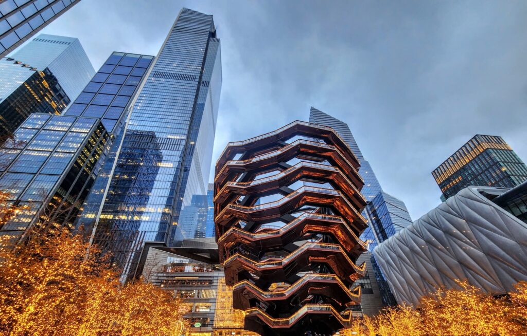 The copper-colored honeycomb staircases of the Vessel at Hudson Yards at dusk, among glass towers and lit trees