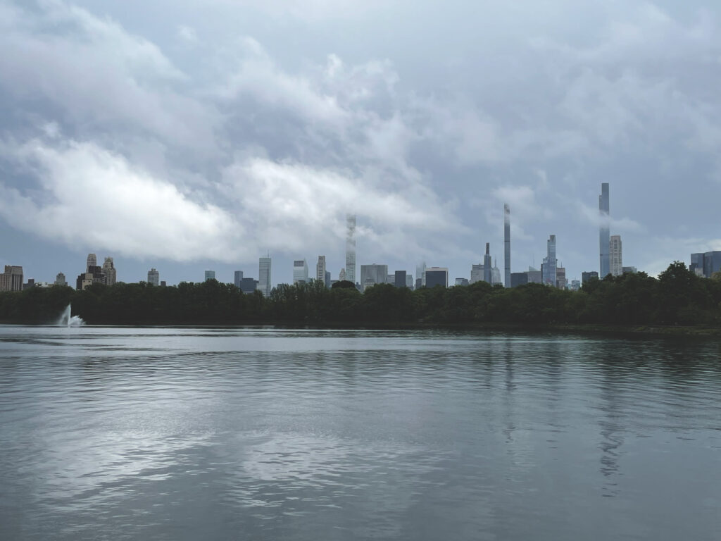 The Central Park Reservoir under a cloudy sky, with the Midtown skyline beyond the trees and its fountain on the left