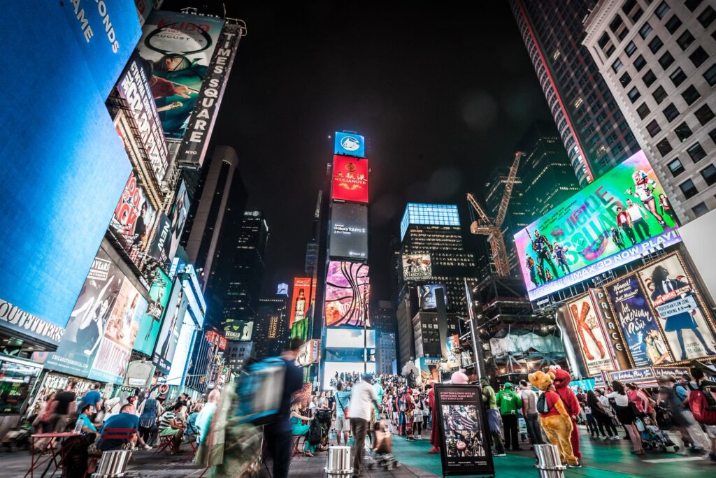 Times Square at night, with towers of lit billboards, Broadway show signs and crowds on the plaza