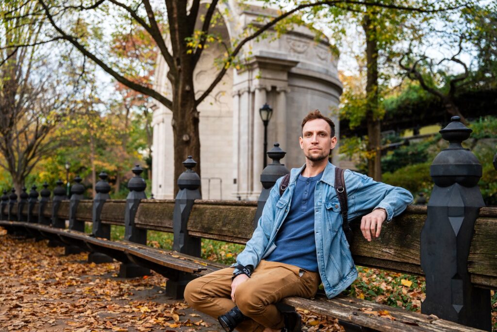 A man sitting on a long wooden bench by the Naumburg Bandshell in Central Park, with fallen leaves on the path