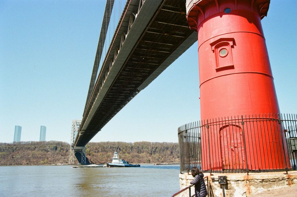 The Little Red Lighthouse on its fenced stone base beneath the George Washington Bridge, with a tugboat on the Hudson River