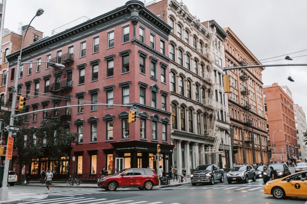 A red-brick corner building and cast-iron facades at a Broome Street crossing in SoHo, with a yellow cab passing