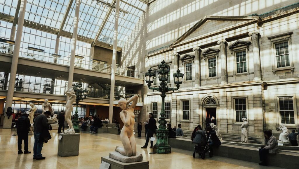 Sunlit glass-roofed sculpture court at the Metropolitan Museum of Art, with marble statues, green lamp posts and a classical stone facade
